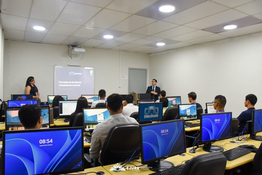 A imagem mostra um laboratório de informática, onde homens e mulheres utilizam computadores. Eles olham para a frente onde há uma mulher à esquerda, e um homem à direita. Ao centro, há um quadro branco com informações projetadas.