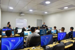 A imagem mostra um laboratório de informática, onde homens e mulheres utilizam computadores. Eles olham para a frente onde há uma mulher à esquerda, e um homem à direita. Ao centro, há um quadro branco com informações projetadas.
