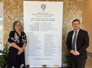 A imagem mostra à esquerda uma mulher de cabelos grisalhos cacheados, que usa óculos, sorri levemente e está de vestido preto com detalhes brancos. No centro, há uma placa branca com letras pretas do Poder Judiciário. Do lado direito, está um homem branco que sorri. Ele usa paletó escuro.