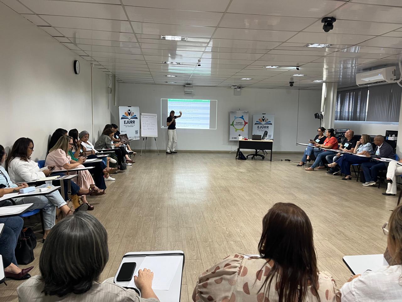 A imagem mostra um auditório em formato semicircular. Diferentes pessoas sentadas observam a formadora que está centro próximo a uma imagem projetada na pareda. Dos lados esquerdo e direito, há banners da Ejurr, um do Programa Justiça Comunitária e um flip chart (conhecido como bloco de cavalete.)