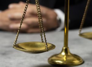 A imagem mostra em destaque a balança dourada da justiça. Ela está sob uma mesa, enquanto um par de mãos estão desfocadas ao fundo, apoiadas no móvel