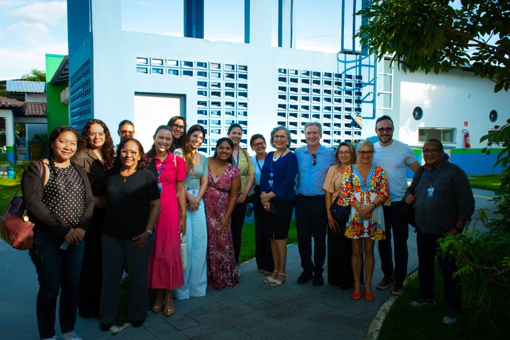 Um grupo de dezesseis pessoas, compostas por homens e mulheres, estão em frente a um prédio de coloração branca. O grupo posa para uma foto, na direção do observador.