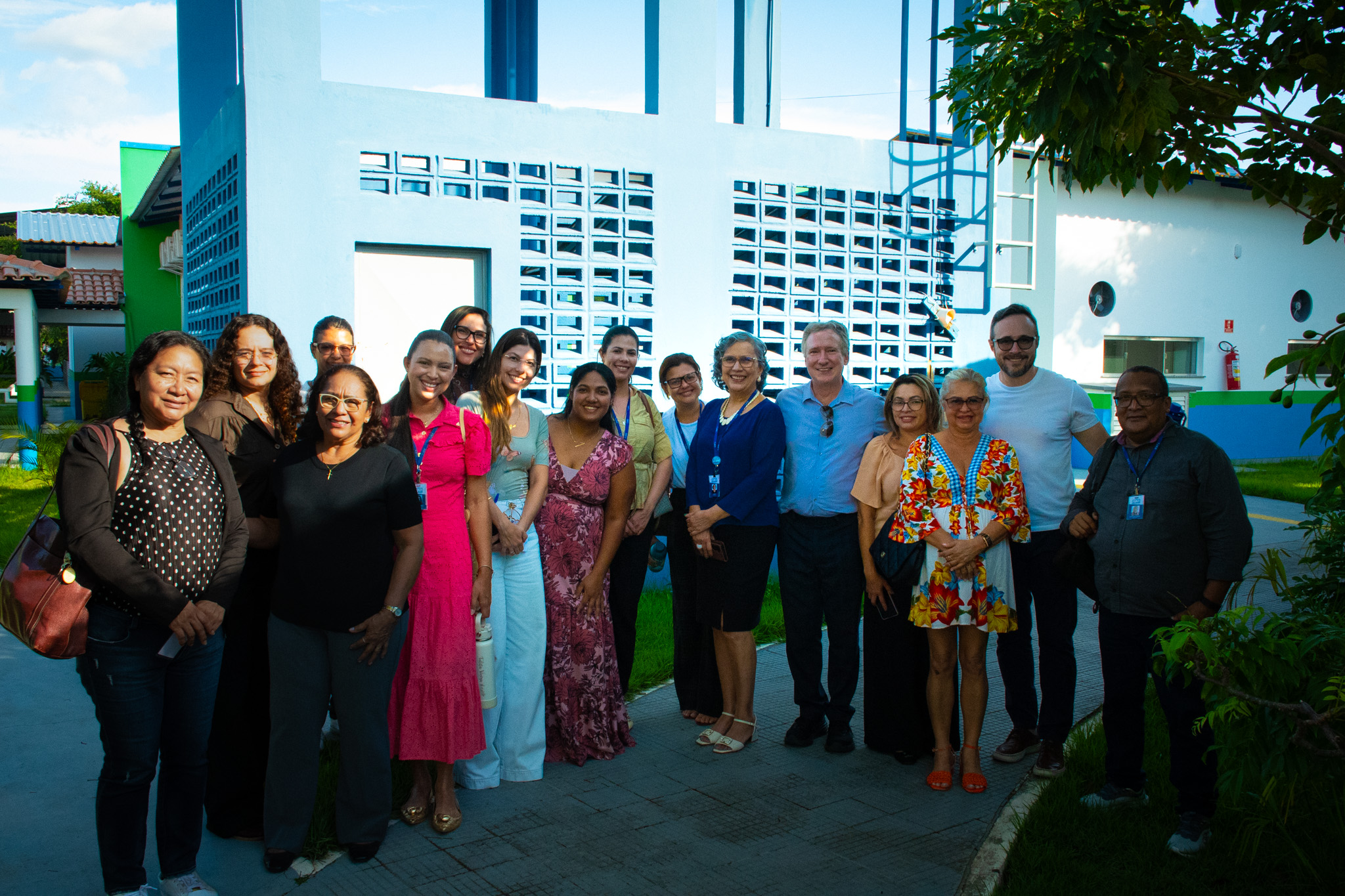 Um grupo de dezesseis pessoas, compostas por homens e mulheres, estão em frente a um prédio de coloração branca. O grupo posa para uma foto, na direção do observador.