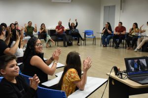 A imagem mostra uma sala de aula com pessoas sentadas em formato circular; Elas se comunicam em Libras através de gestos com as mãos.