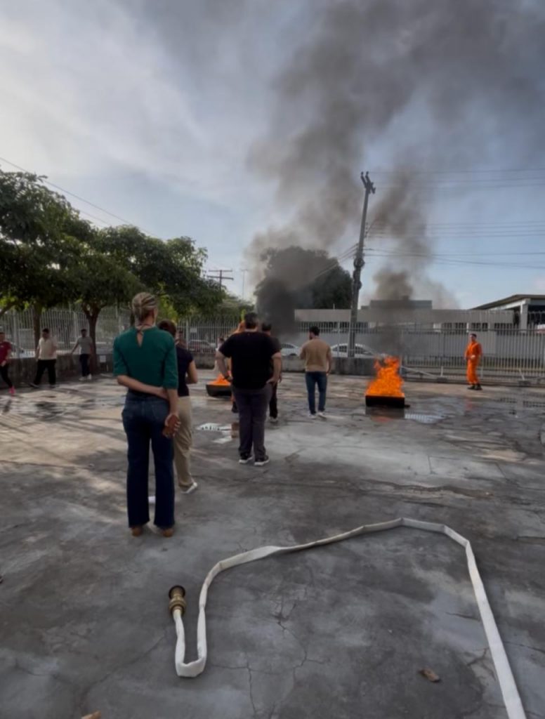 A imagem mostra pessoas de pé, em fila e de costas, tendo ao lado uma mangueira de água clara. Na parte da frente, há uma pequena fogueira com uma fumaça escura, e um bombeiro militar próximo conversando.