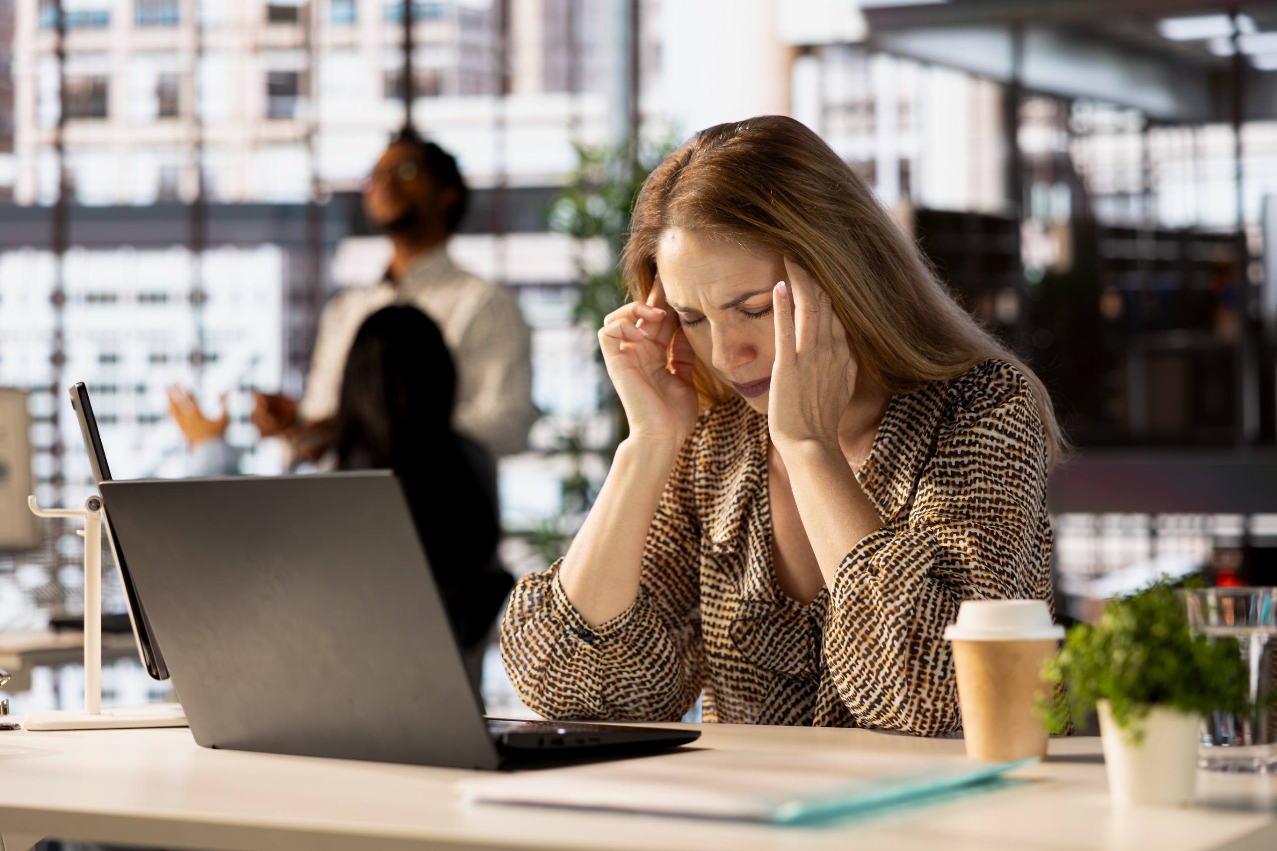 A imagem mostra uma mulher de cabelos claros com as mãos nas laterais da cabeça, com expressão séria e olhos fechados. Ela está de frente para um notebook spbre uma mesa. Ao fundo, pessoas conversam e a imagem está levemente desfocada.