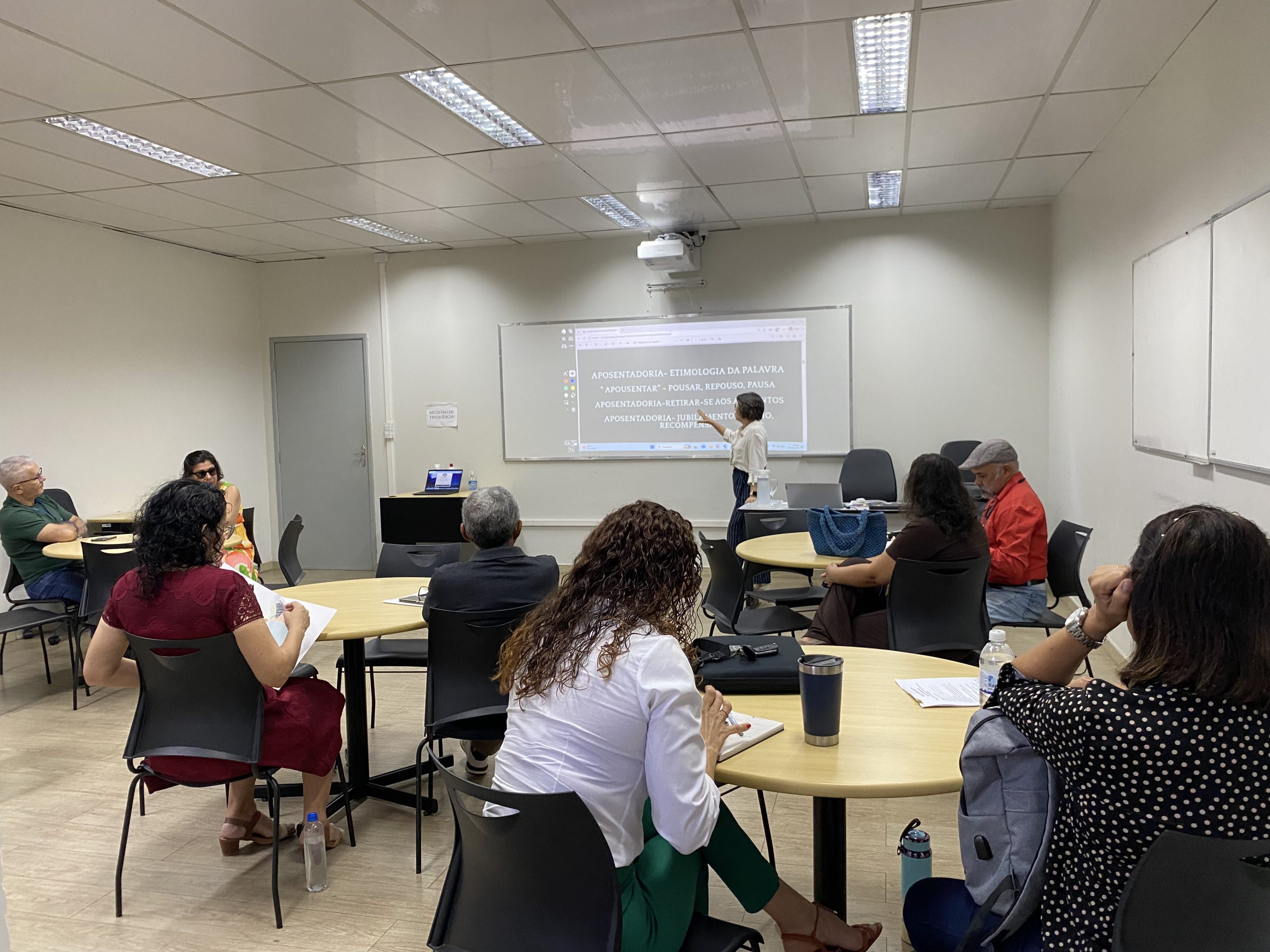 A matériam possui três imagens, sendo a foto da capa, a terceira imagem. A seguir a descrição na ordem de publicação no texto:  Foto 1:  Fotografia em close de duas apostilas brancas sobre uma mesa de madeira. Na capa, veem-se o logotipo "Geronto Ser - Ações para o Envelhecimento Saudável", o nome da facilitadora Maria Tereza Sampaio de Carvalho e o título "Workshop Para Além da Aposentadoria", acompanhados por um desenho estilizado de uma árvore com raízes aparentes. Na apostila em primeiro plano, lê-se o nome "Elizângela" escrito à mão no campo reservado. Foto 2: Fotografia de uma sala de aula iluminada onde ocorre o Workshop "Para Além da Aposentadoria". Vários participantes estão sentados ao redor de mesas redondas distribuídas pelo ambiente com piso de madeira. À esquerda, uma palestrante de blusa branca e calça listrada gesticula enquanto conduz a apresentação para o grupo. Foto 3: Outro ângulo da sala durante o Workshop "Para Além da Aposentadoria". A facilitadora está de pé em frente a um telhado de projeção exibindo slides com o texto "Aposentadoria - Etimologia da Palavra". Os participantes acompanham a palestra sentados em mesas redondas, com cadernos e anotações.