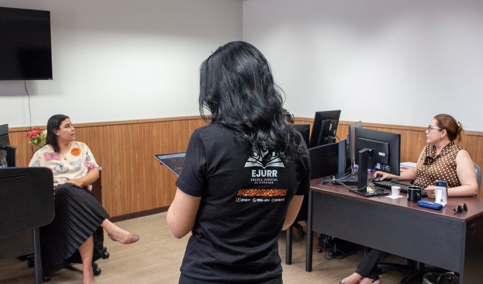 A imagem mostra uma mulher de costas com camisa preta onde está a logo da Ejurr. Em cada lateral, há uma mesa com computador onde estão sentadas duas mulheres.
