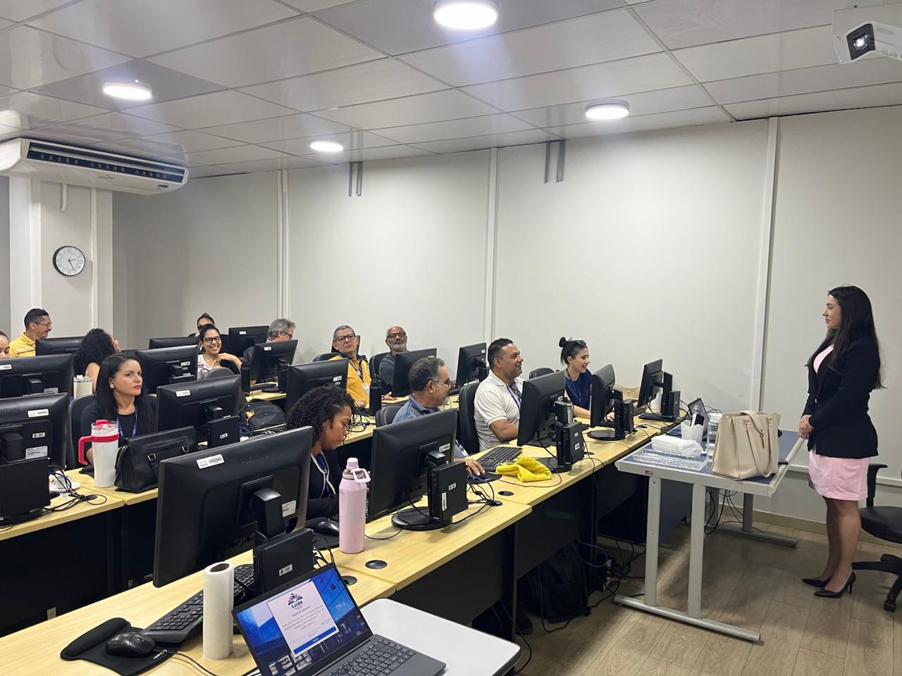 A imagem mostra uma mulher de blazer preto e vestido rosa em frente de um grupo sentado em um laboratório de informática.