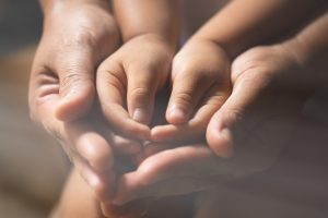 A imagem mostra quatro mãos juntas , sendo um adulto e uma criança. As mãos da crianaça estão por cima da mão adulta lembrando um coração.