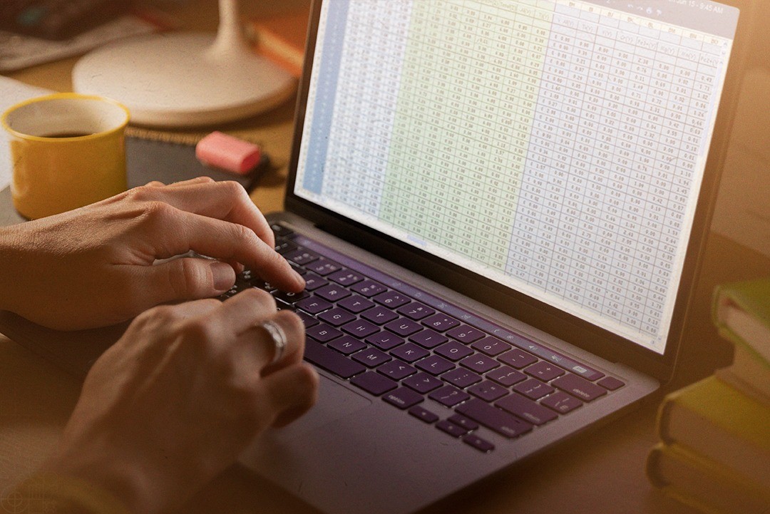 Em um notebook com planilhas numéricas, uma pessoa digita no teclado. O equipamento está sobre uma mesa com uma xícara e objetos que remetem a um ambiente de escritório