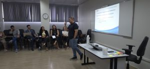 Em um ambiente de sala de aula, o instrutor está de pé, fala e gesticula. Ele é um homem pardo, com traços orientais e calvo, veste blusa escura e calça jeans,, estando à frente de uma imagem projetada em um quadro de sala de aula branco. À esquerda, está uma mesa com notebook e objetos diversos. Do lado direito, várias pessoas sentadas observam, entre homens e mulheres.