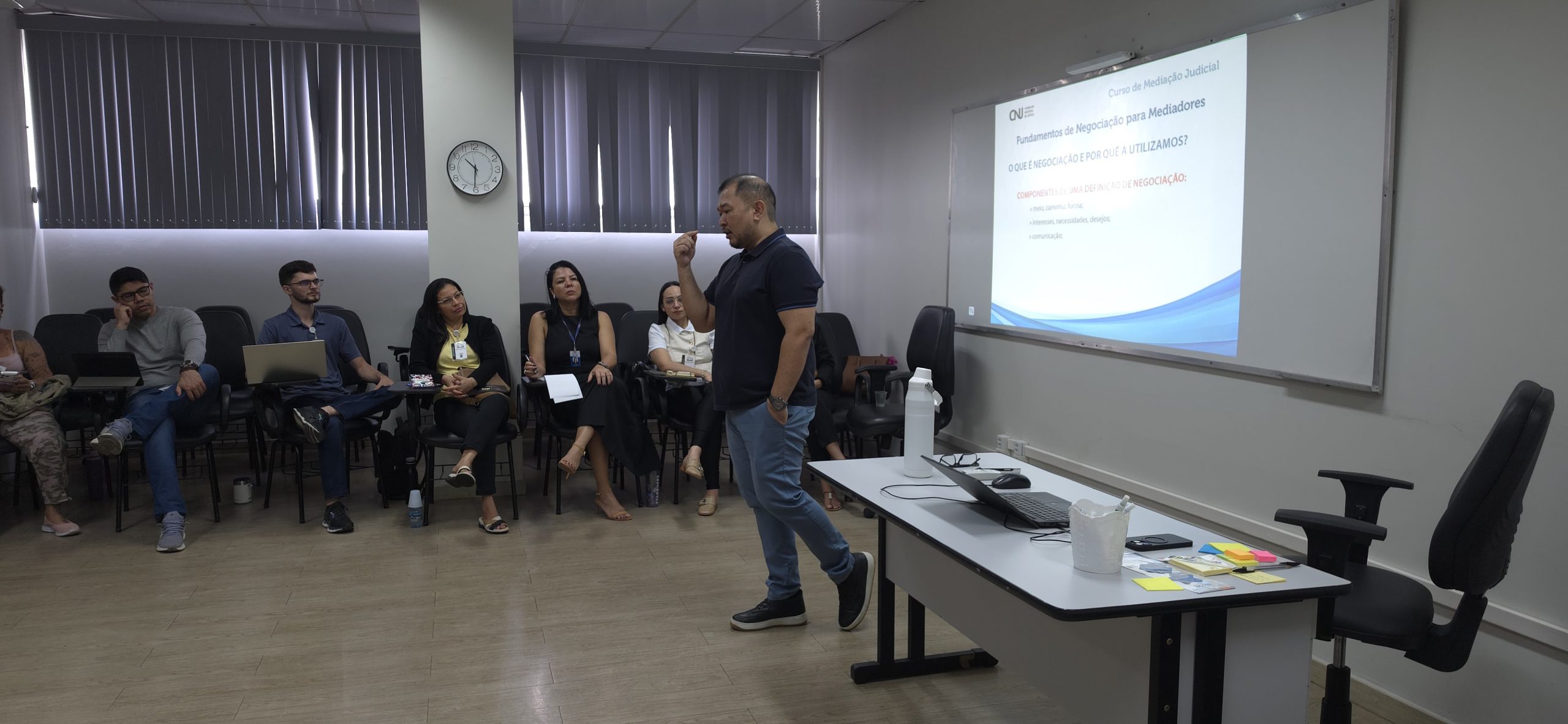 Em um ambiente de sala de aula, o instrutor está de pé, fala e gesticula. Ele é um homem pardo, com traços orientais e calvo, veste blusa escura e calça jeans,, estando à frente de uma imagem projetada em um quadro de sala de aula branco. À esquerda, está uma mesa com notebook e objetos diversos. Do lado direito, várias pessoas sentadas observam, entre homens e mulheres.