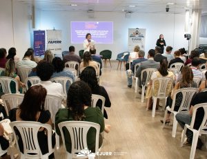 A imagem mostra um auditório onde há pessoas sentadas e de costas em cadeiras brancas de plástico. Uma mulher fala com o microfone enquanto segura um texto. Atrás dele há uma projeção na parede. Nas laterais, há banners e uma intérprete de Libras.