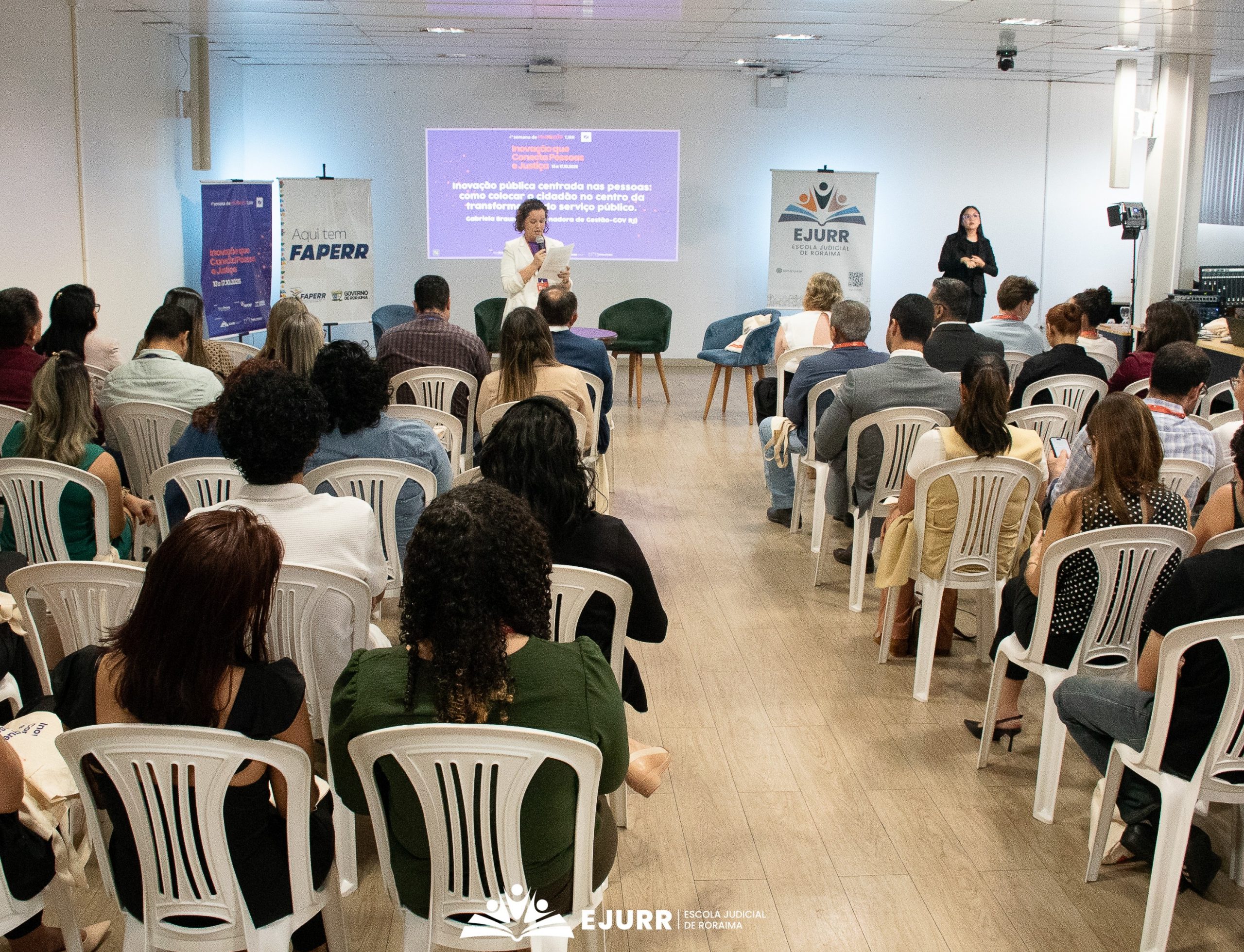 A imagem mostra um auditório onde há pessoas sentadas e de costas em cadeiras brancas de plástico. Uma mulher fala com o microfone enquanto segura um texto. Atrás dele há uma projeção na parede. Nas laterais, há banners e uma intérprete de Libras.