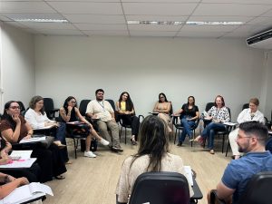 A imagem mostra um grupo de pessoas, entre homens e mulheres, sentados em formato circular em uma sala com paredes claras. Alguns estão de costas e outros observam os formadores.