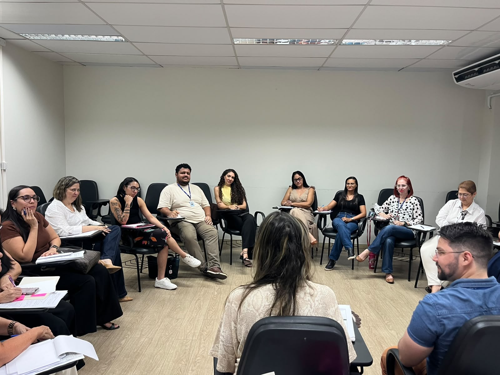 A imagem mostra um grupo de pessoas, entre homens e mulheres, sentados em formato circular em uma sala com paredes claras. Alguns estão de costas e outros observam os formadores.