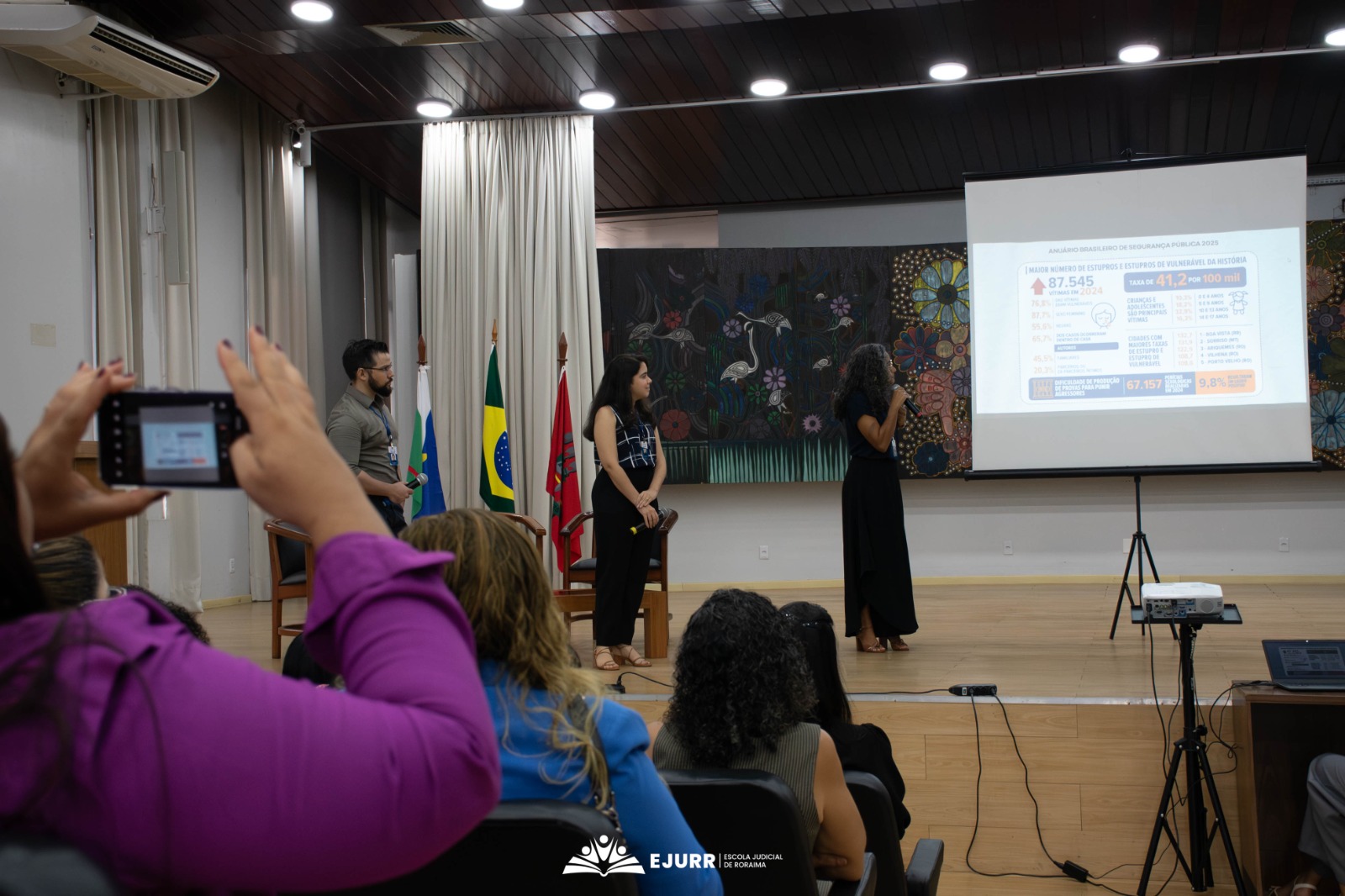 A imagem mostra um auditório bem iluminado, com uma decoração composta por três bandeiras, um quadro e cortinas brancas. No ambiente, há uma tela exibindo uma série de dados pelo datashow. No local, sete pessoas observam o que está sendo mostrado pela tela, sendo quatro delas parte do público e três em cima do palco.