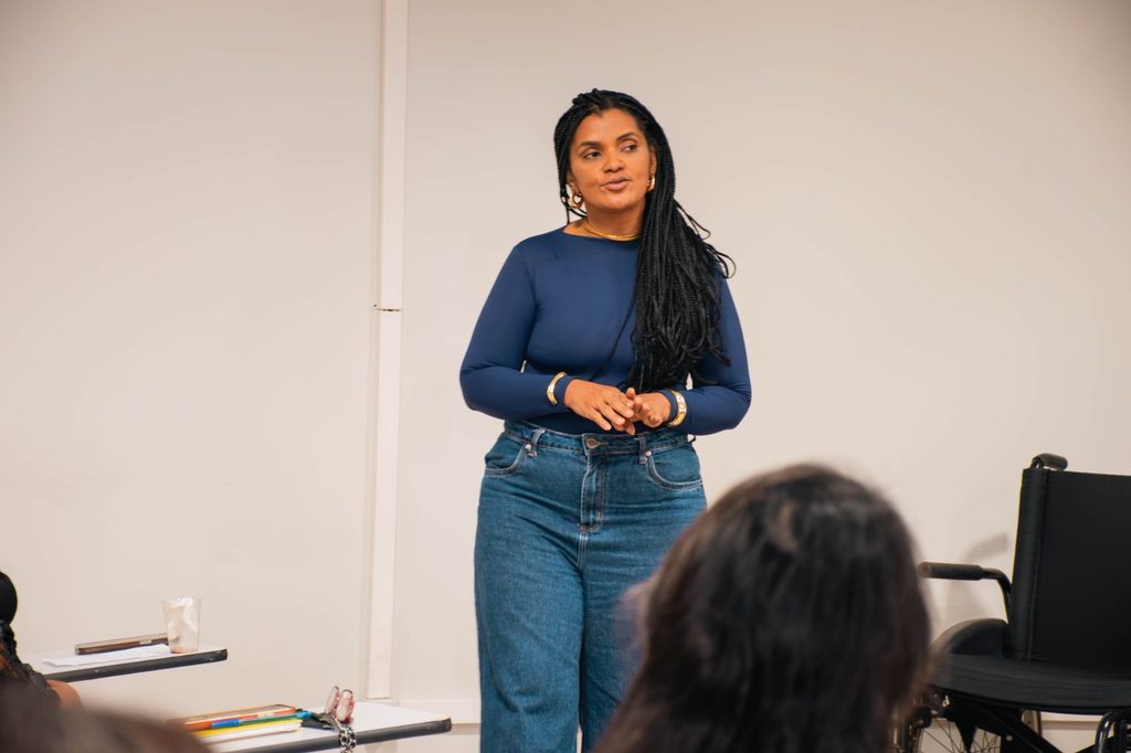 A imagem mostra uma mulher negra, vestida com uma blusa azul escura e uma calça jeans azul em uma sala de aula. Ela está de pé, com as mãos em frente ao corpo, enquanto olha para o lado.