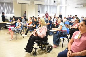 A imagem mostra um grupo de pessoas participando de uma palestra ou capacitação em uma sala de aula, com cadeiras organizadas em fileiras e equipamentos de gravação. Entre os participantes, há uma pessoa utilizando cadeira de rodas, acompanhando a atividade.