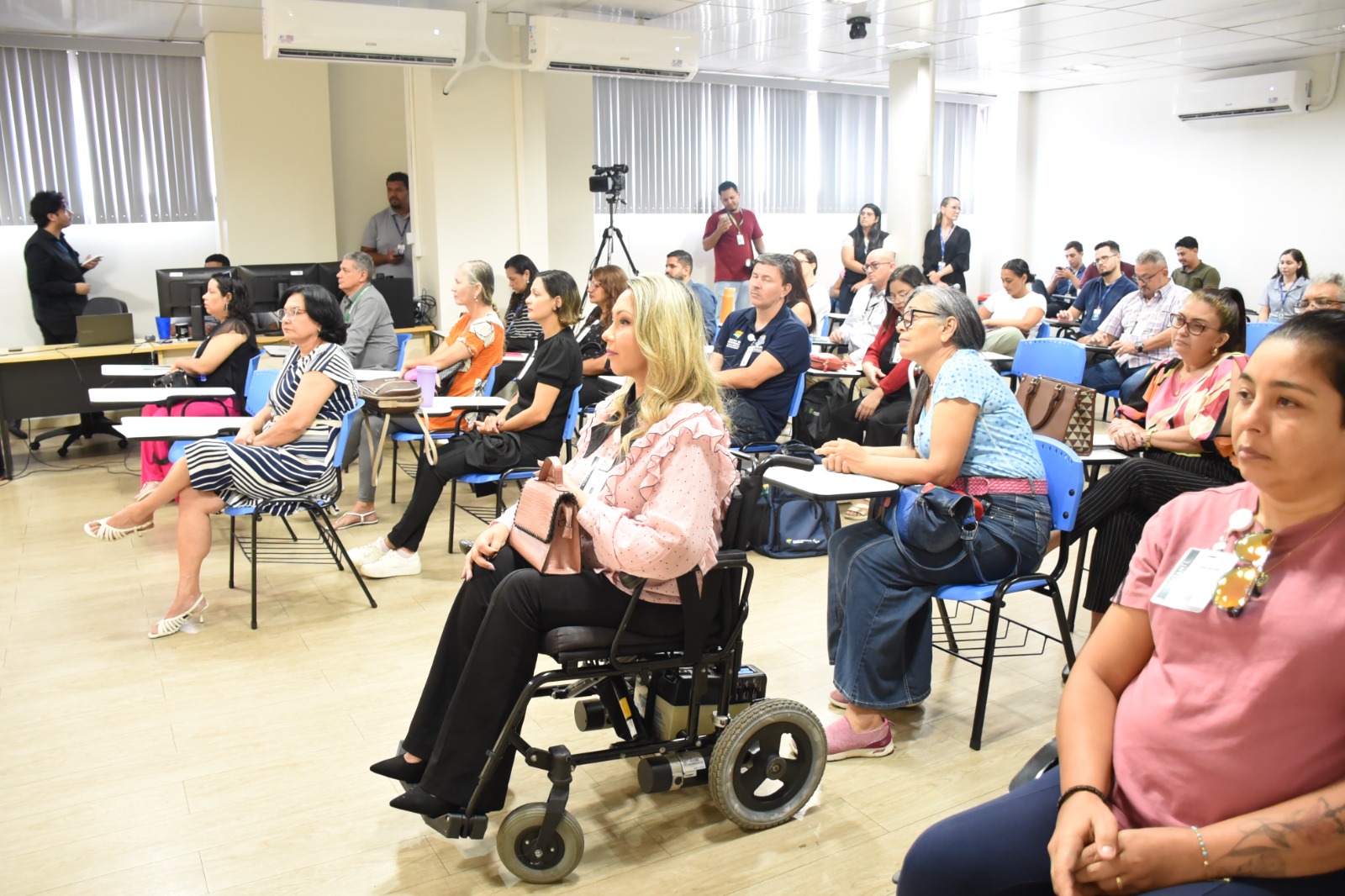 A imagem mostra um grupo de pessoas participando de uma palestra ou capacitação em uma sala de aula, com cadeiras organizadas em fileiras e equipamentos de gravação. Entre os participantes, há uma pessoa utilizando cadeira de rodas, acompanhando a atividade.