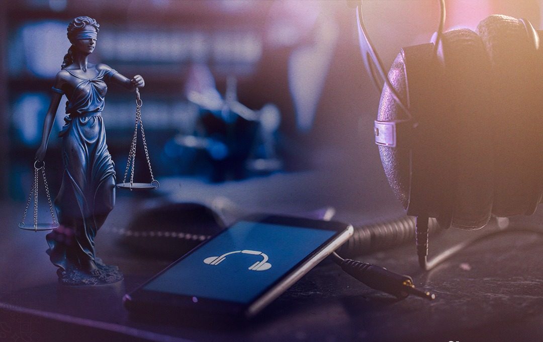 A imagem mostra uma mesa com três objetos: uma estátua da figura de Têmis, um dispositivo eletrônico mostrando uma tela azul com um ícone branco de fone de ouvido e, na mesa, há também um fone de ouvido. Ao fundo, está uma estante com livros e a figura parcial de uma pessoa, ambos desfocados