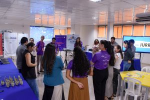 A imagem mostra pessoas em formato circular, entre homens e mulheres, sendo que todos estão em pé. Elas estão um ambiente com cadeiras e mesas coloridas, com grandes monitores que expõem frases e instituições diversas, há ainda banners da Ejurr, da Semana da Inovação e do Roraima Inova.