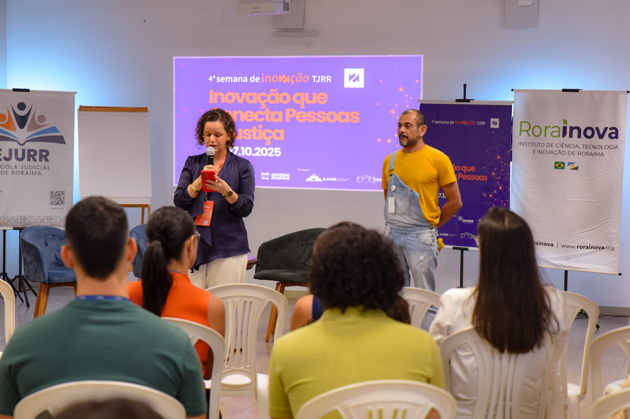Um homem e uma muher estão de frente para o público. NO fundo, a projeção da Semana da Ibnovaçãoç CAD
