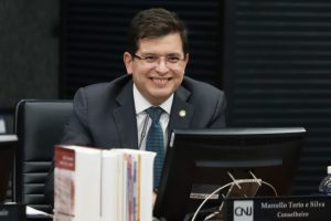 A imagem mostra um homem branco, de óculos, cabelo castanho eque sorri. Ele usa paletó e gravata com quadriculados pequenos na cor verde petróleo. Na sua frente, está um monitor e livros em pé. O fundo é escuro e aparece desfocado.