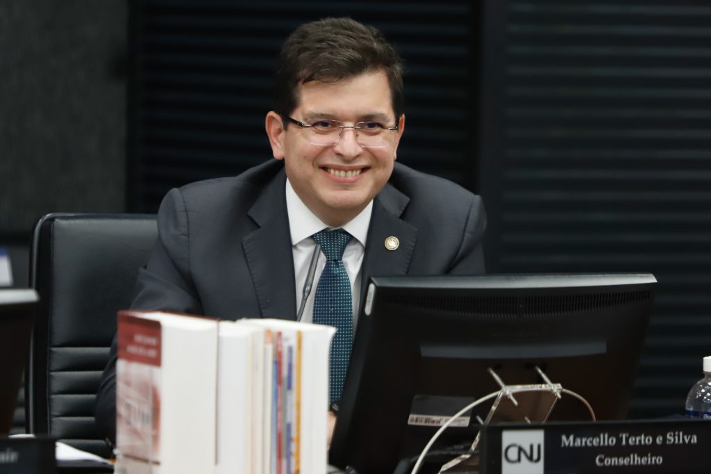 A imagem mostra um homem branco, de óculos, cabelo castanho eque sorri. Ele usa paletó e gravata com quadriculados pequenos na cor verde petróleo. Na sua frente, está um monitor e livros em pé. O fundo é escuro e aparece desfocado.