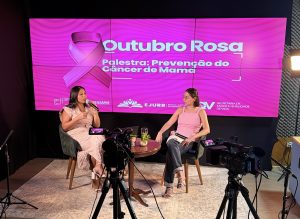Imagem colorida mostra duas mulheres sentadas em poltronas lado a lado, durante uma transmissão ao vivo. Atrás delas, há um telão com o texto: “OUTUBRO ROSA – Palestra: Prevenção do Câncer de Mama”. À frente das mulheres, duas câmeras estão posicionadas em sua direção para captar a transmissão ao vivo.