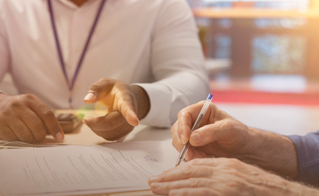 Em destaque estão mãos e um documento, onde uma pessoa assina e a outra aponta em direção ao que está sendo assinado.