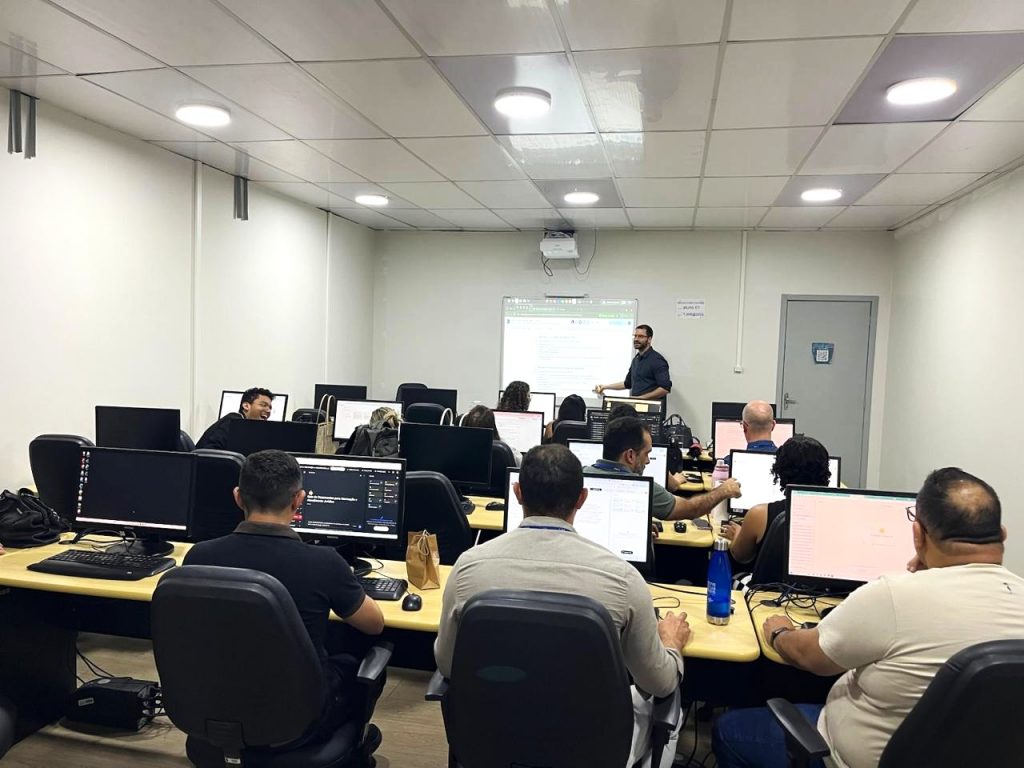  A imagem mostra um ambiente de sala que mestra sala de aula e laboratório de informática, onde pessoas estão sentadas e algumas conversam entre si. À frente da sala, há um homem de cabelo  curto escuro, barba e óculos,blusa preta, que aponta para o quadro branco.