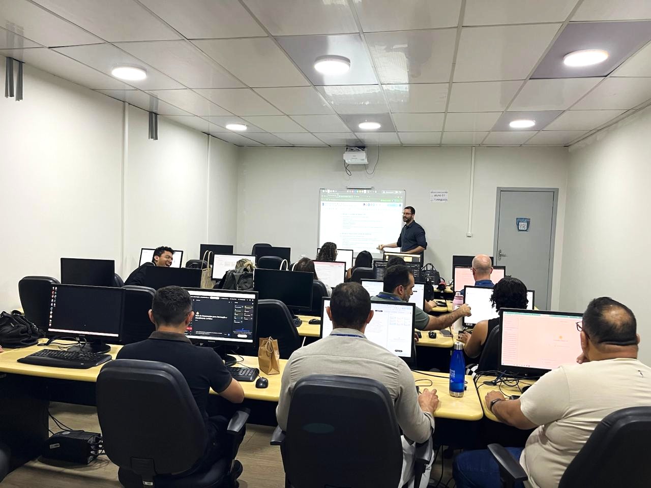 A imagem mostra um ambiente de sala que mestra sala de aula e laboratório de informática, onde pessoas estão sentadas e algumas conversam entre si. À frente, um homem de cabelo baixo escuro, barba e óculos, vestindo blusa preta. Ele aponta para o quadro branco.